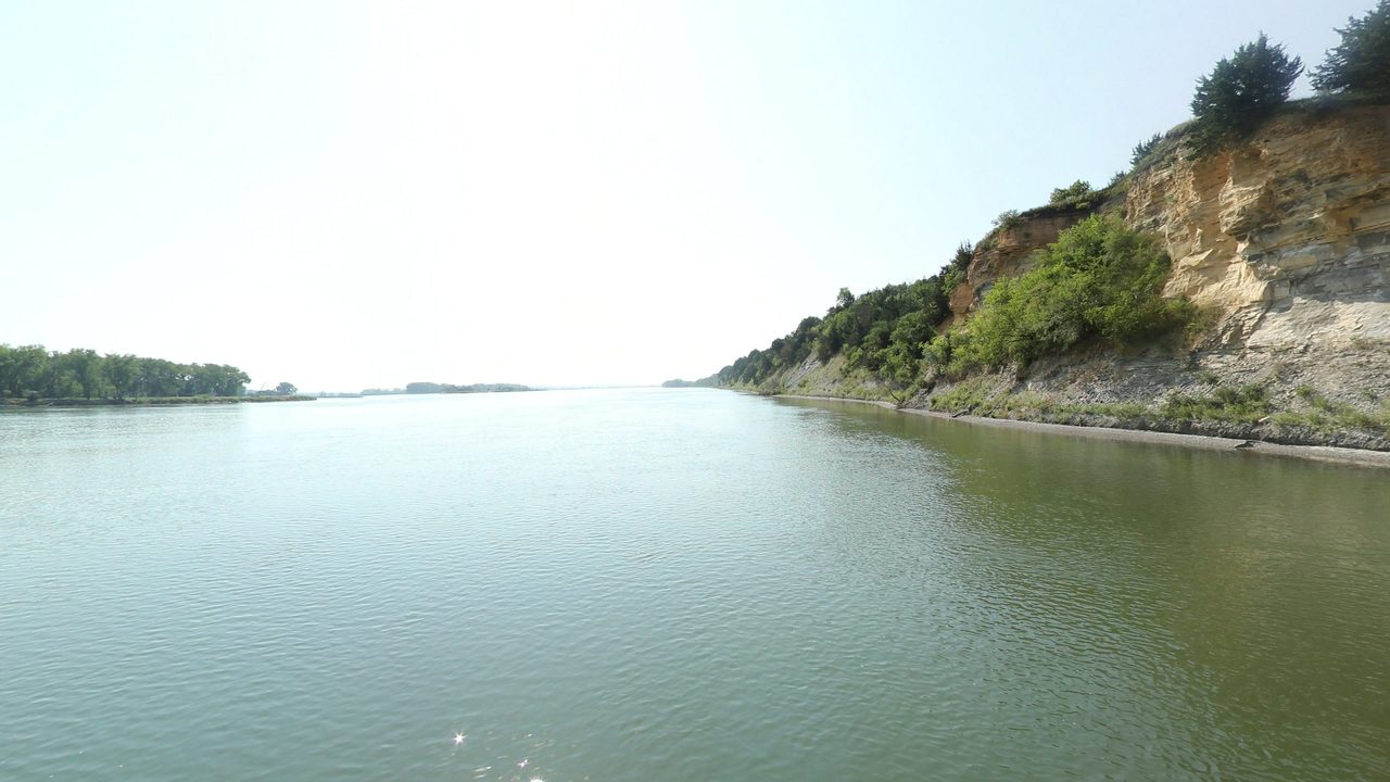 The Missouri River below a pale, eroding bluff topped with trees on the right, 2021