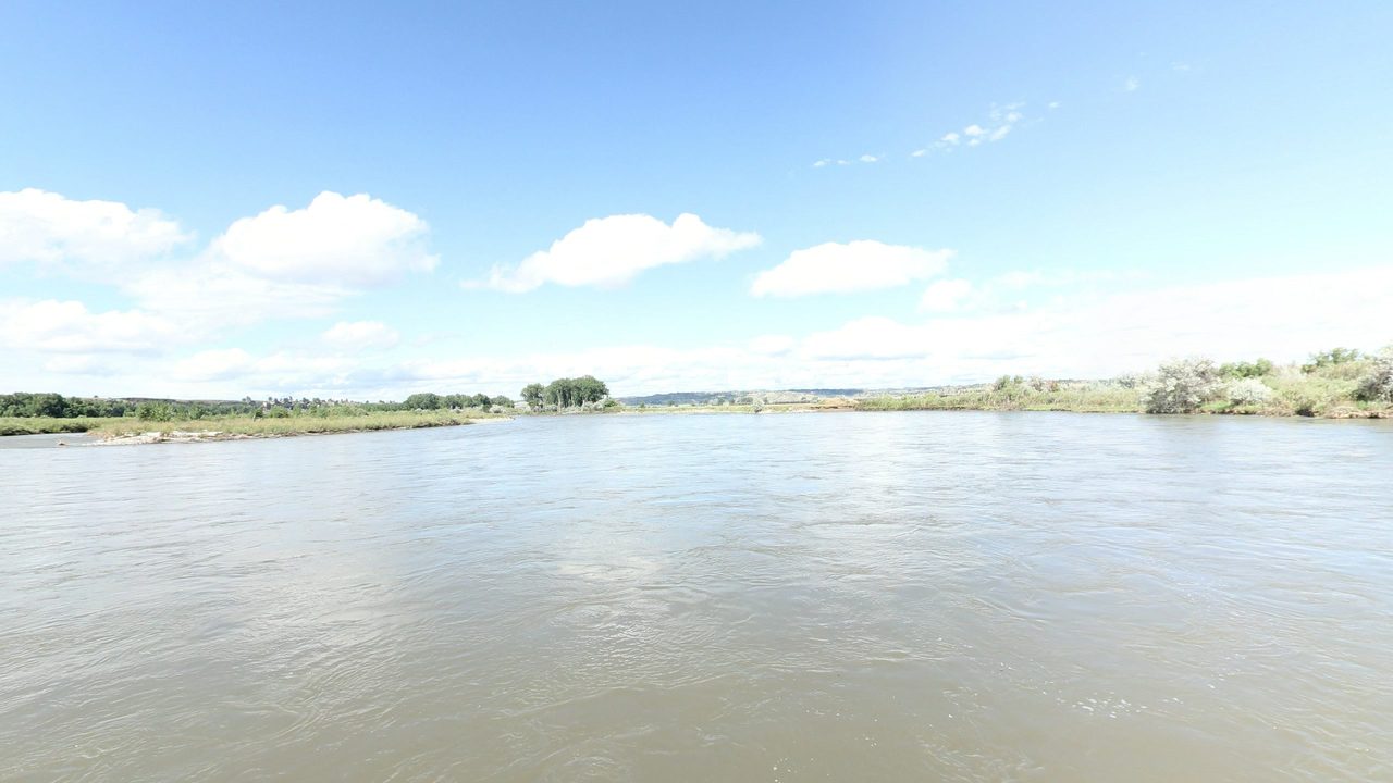 The broad Yellowstone River under white clouds, with low grassy banks and cottonwoods, 2024