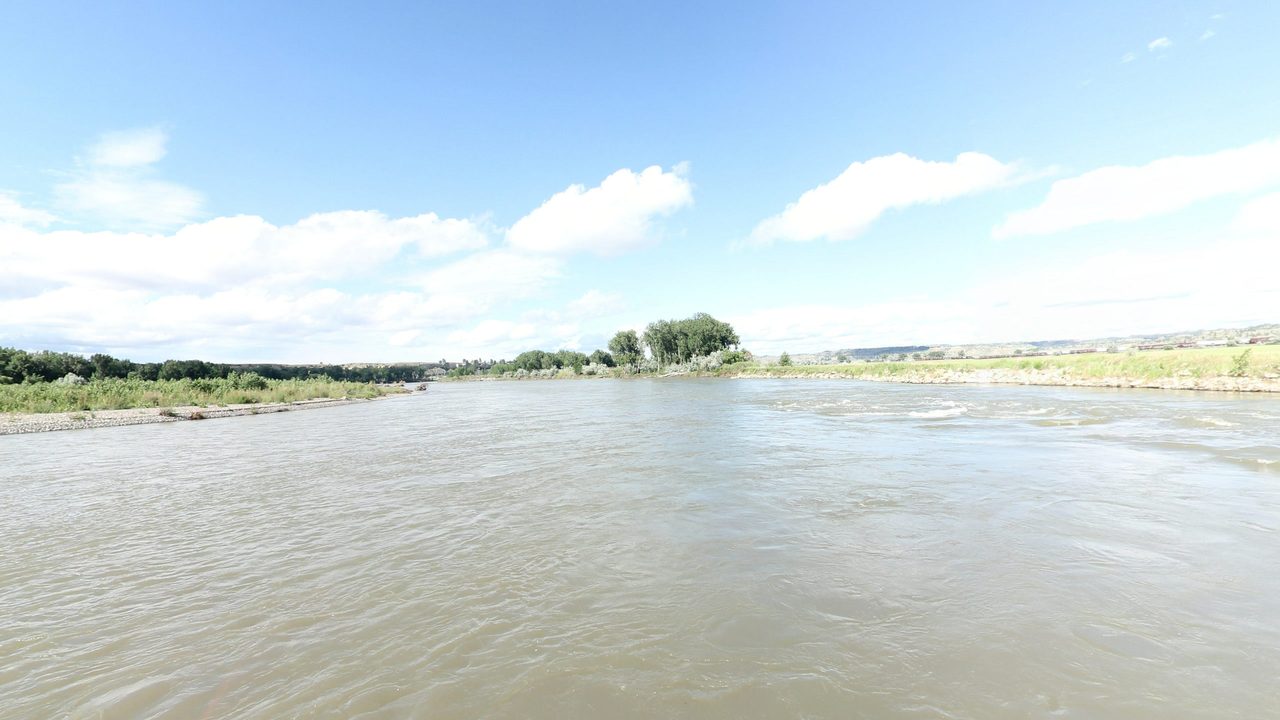 The broad Yellowstone River between low grassy banks under white clouds, 2024