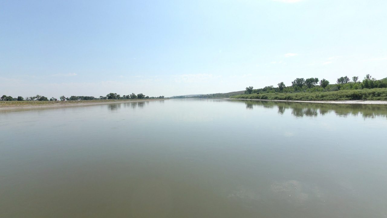 The calm Yellowstone River between low, tree-dotted banks under a hazy sky, 2024