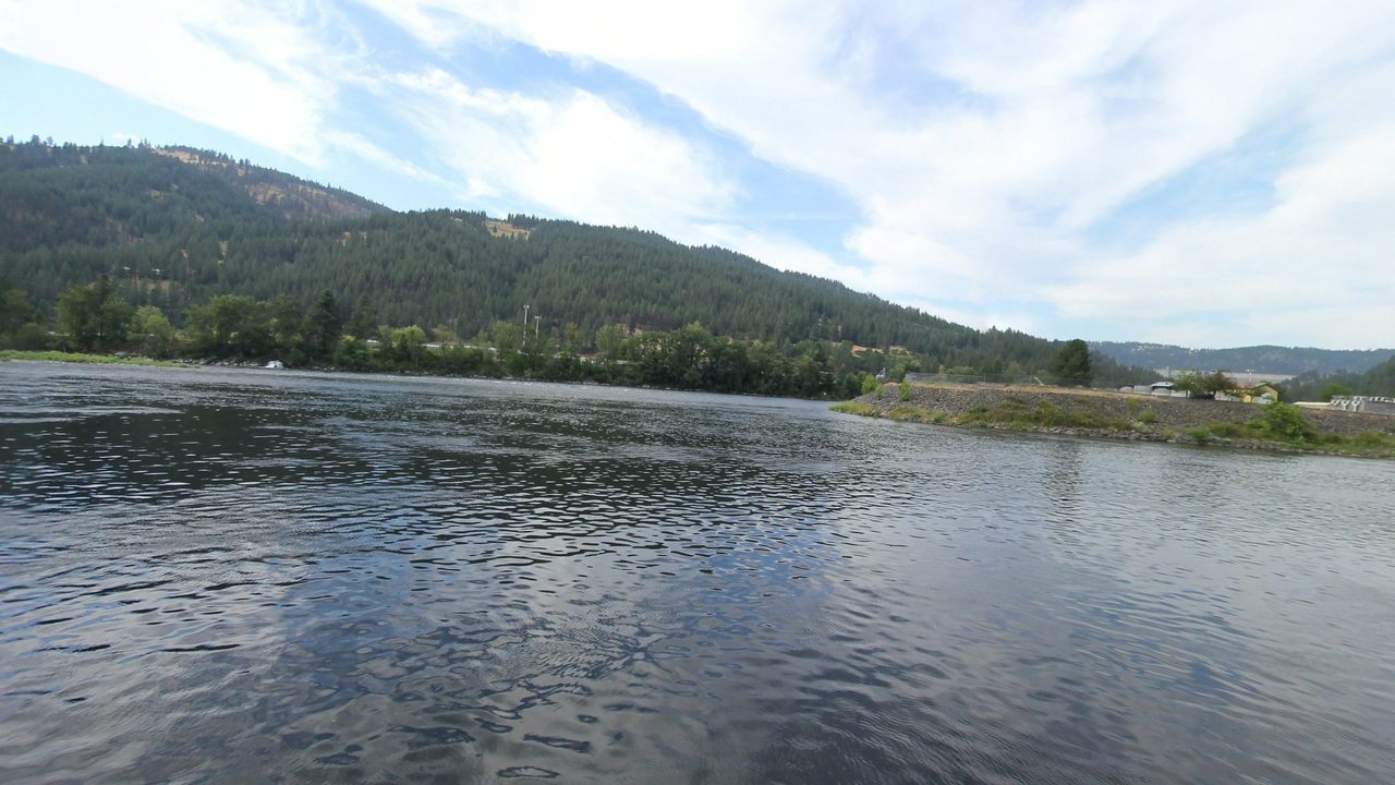 The Clearwater River beside a rock dike, with low industrial buildings and a concrete dam in the distance, 2024