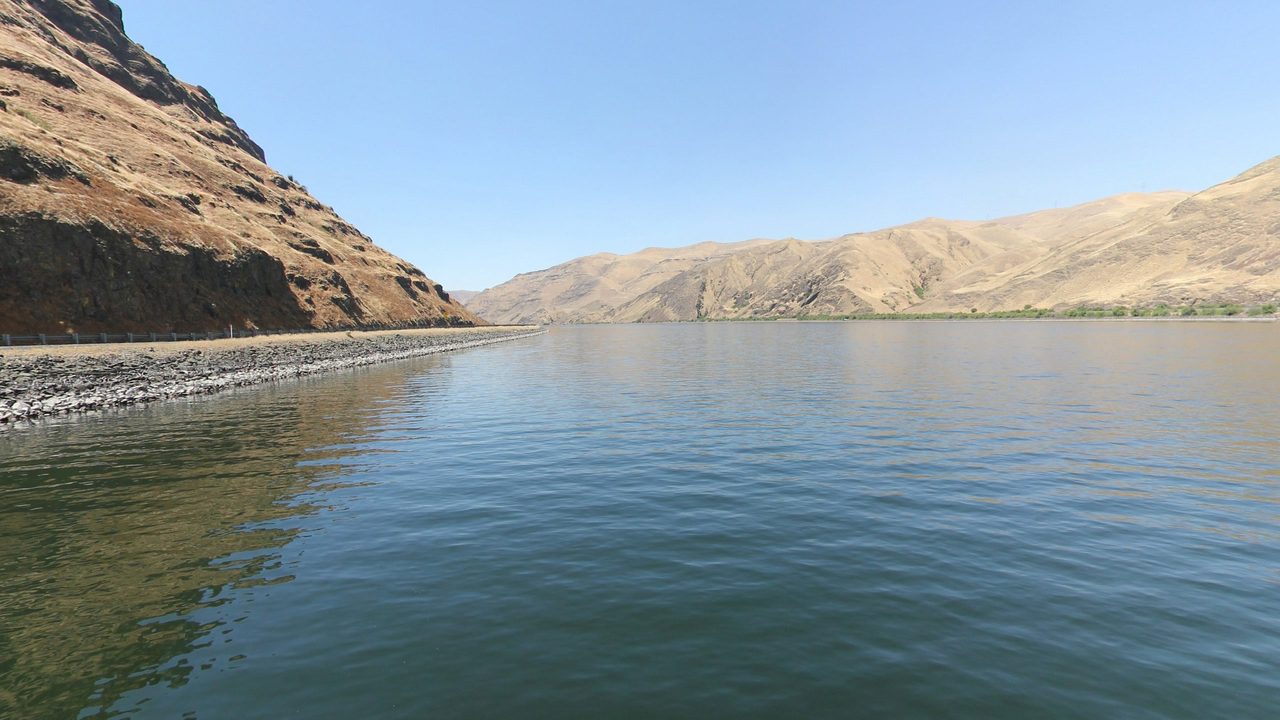 The calm Snake River below a steep, dry cliff, with a guardrailed road on a rocky embankment along the left shore, 2024