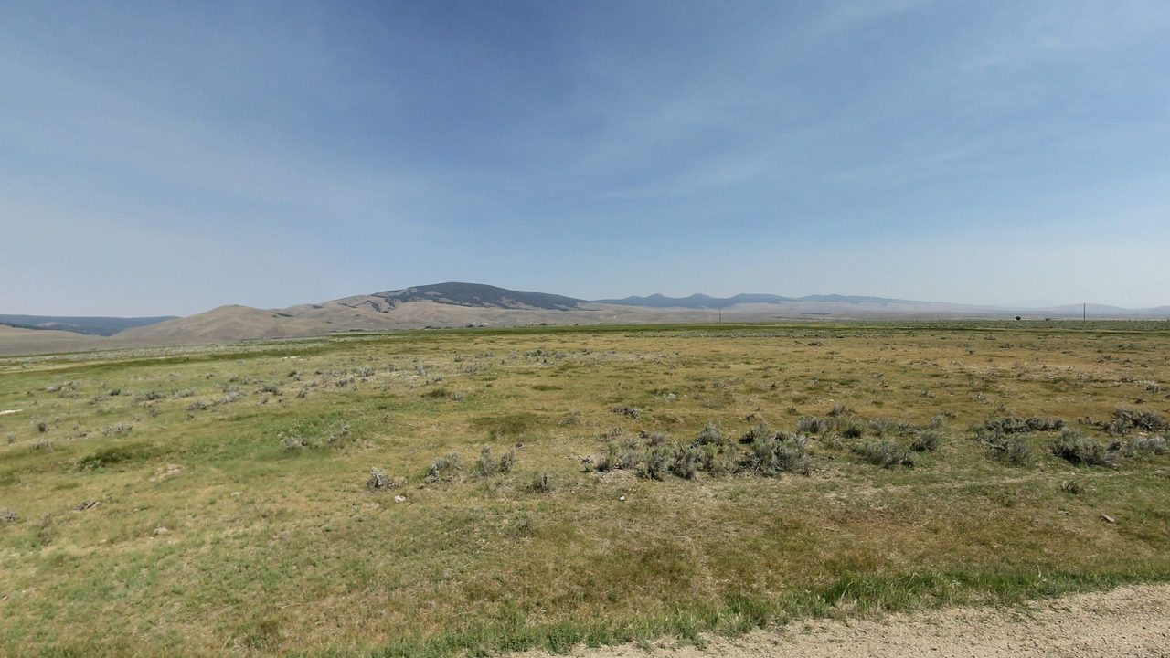A sagebrush pasture stretching to dry, rolling hills and a forested peak under a hazy sky, 2024