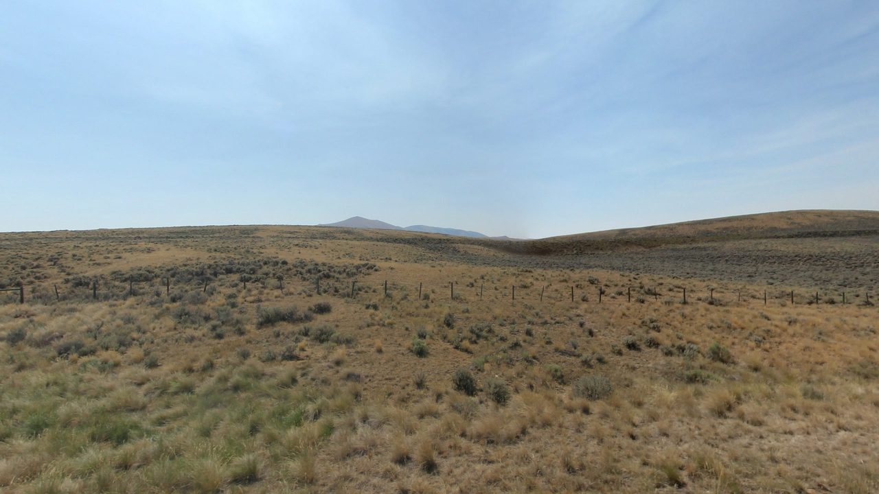 Rolling sagebrush hills behind a barbed-wire fence, with a distant peak under a pale sky, 2024