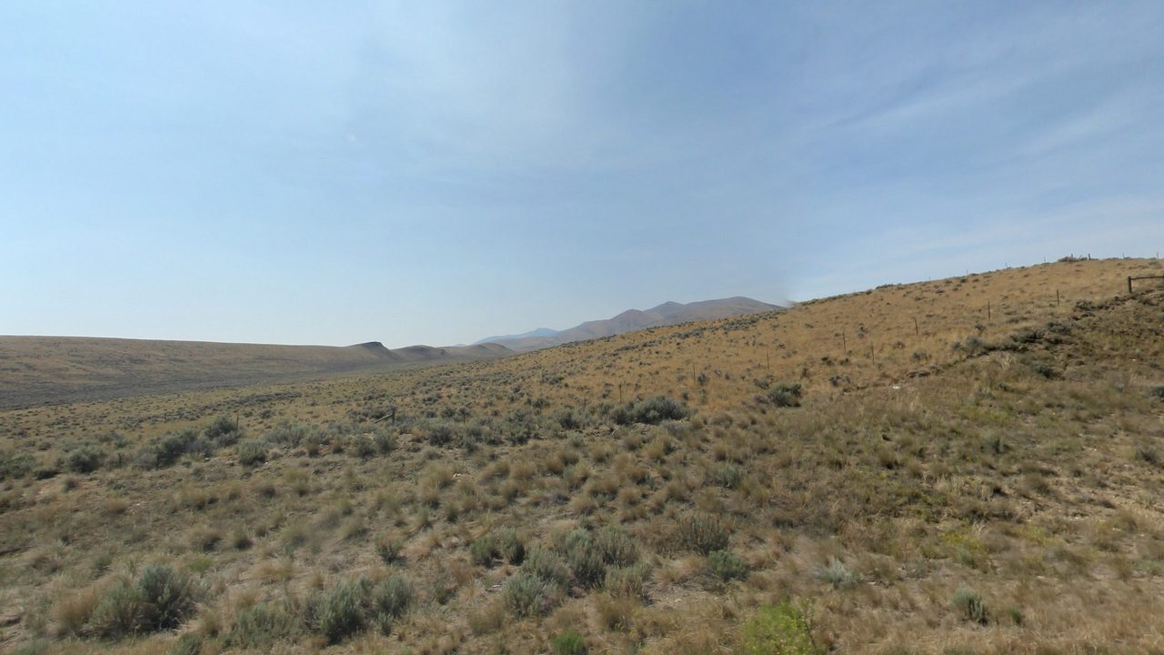 A dry, sagebrush-covered slope rising toward distant mountains under a hazy sky, 2024