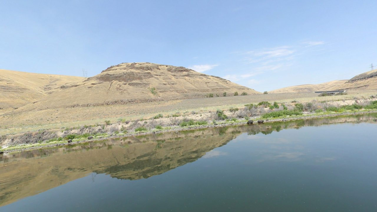 A calm reservoir reflecting dry, grassy hills, with a small steel bridge across a side draw, 2024