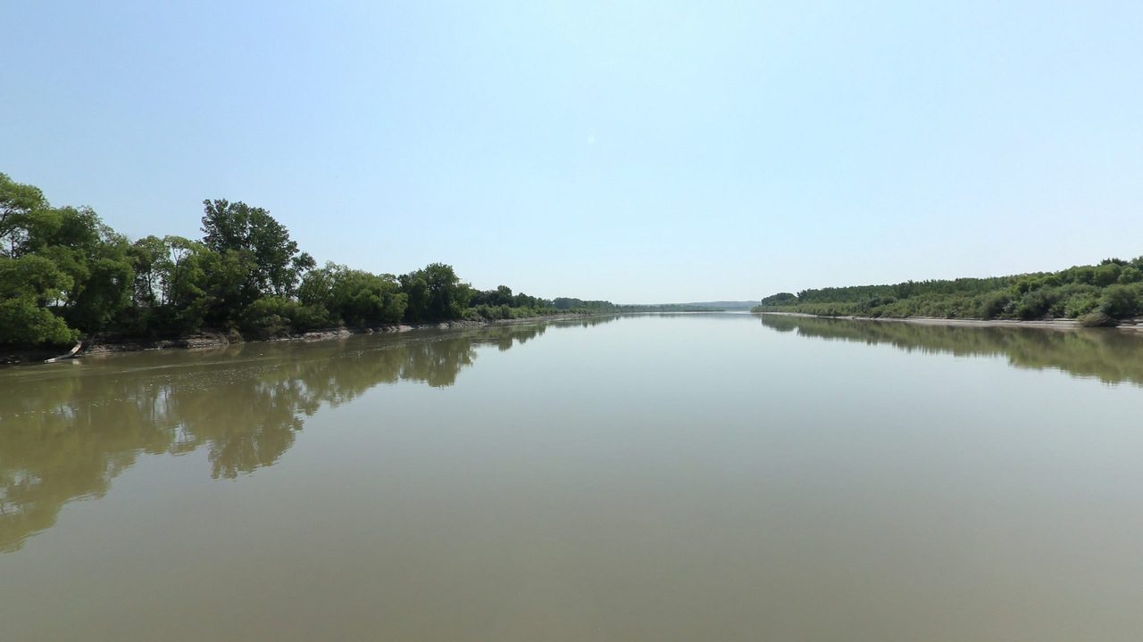 The calm Missouri River reflecting leafy trees on the left bank, under a blue sky, 2024