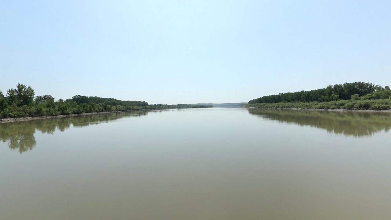 The calm Missouri River between low wooded banks under a clear sky, 2024