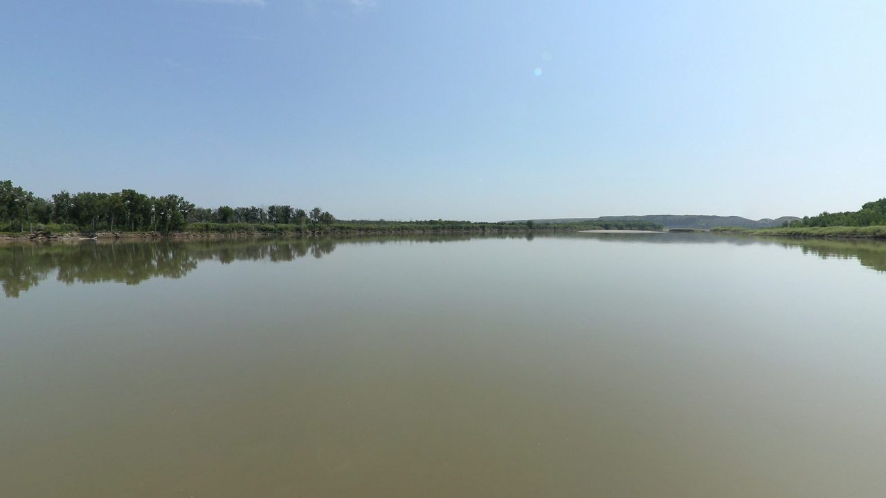 The calm Missouri River reflecting a wooded left bank, with low hills in the distance, 2024