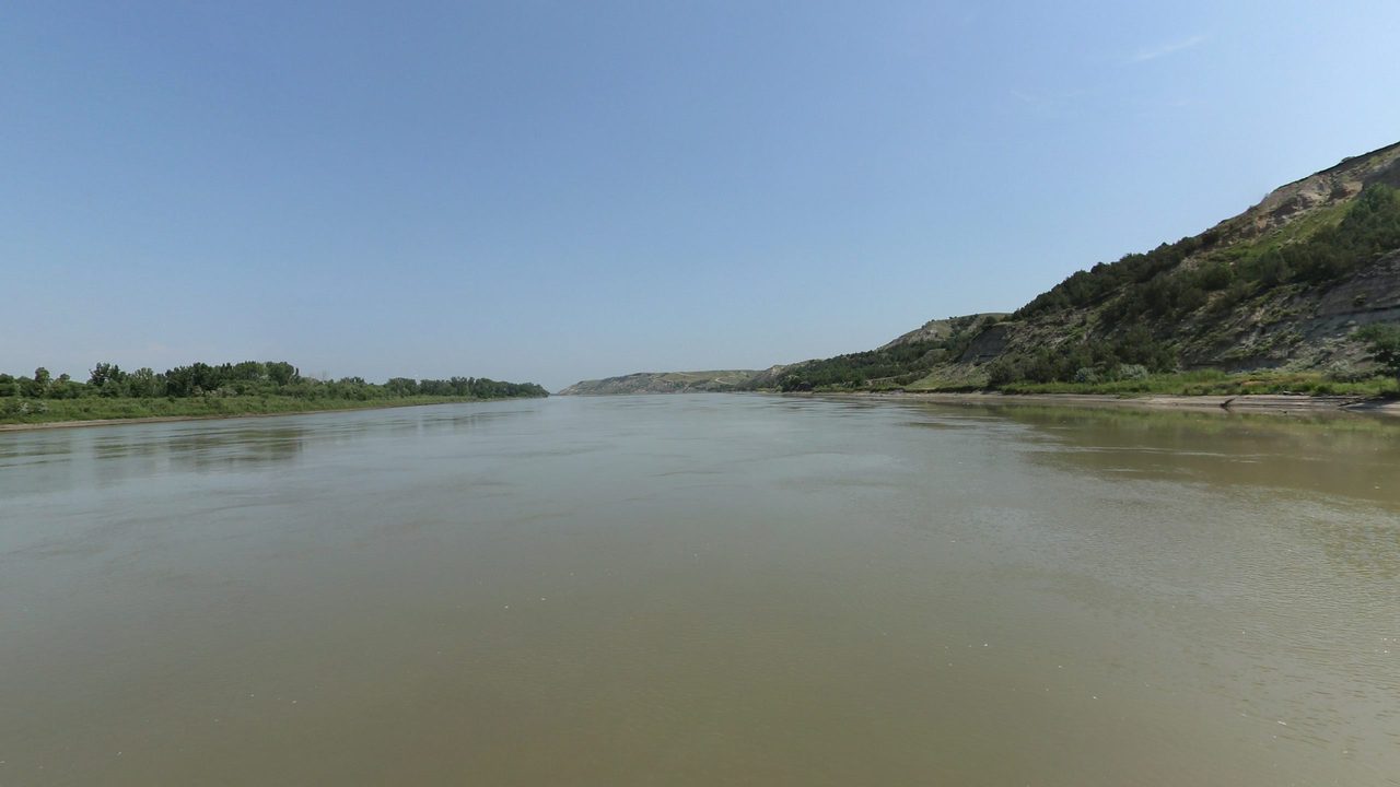 The Missouri River below a steep grassy bluff on the right, under a pale blue sky, 2024