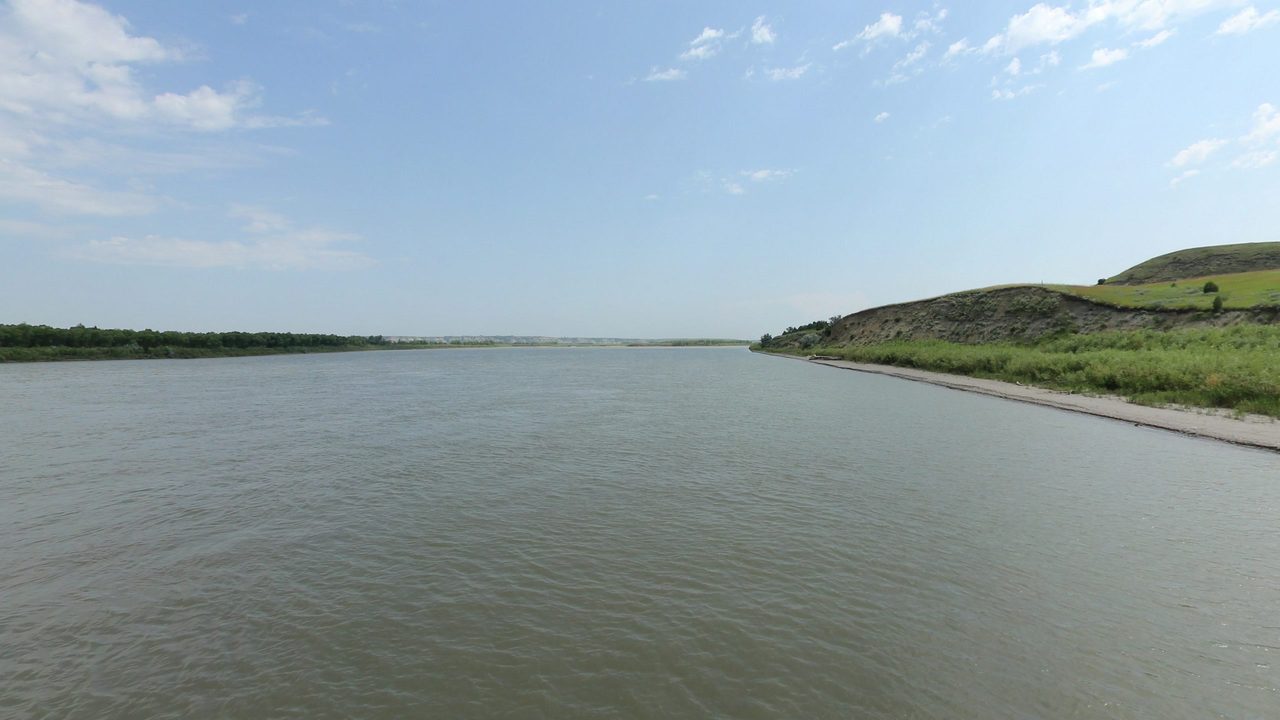 The Missouri River beside a green hill and a sandy shore on the right, under scattered clouds, 2024