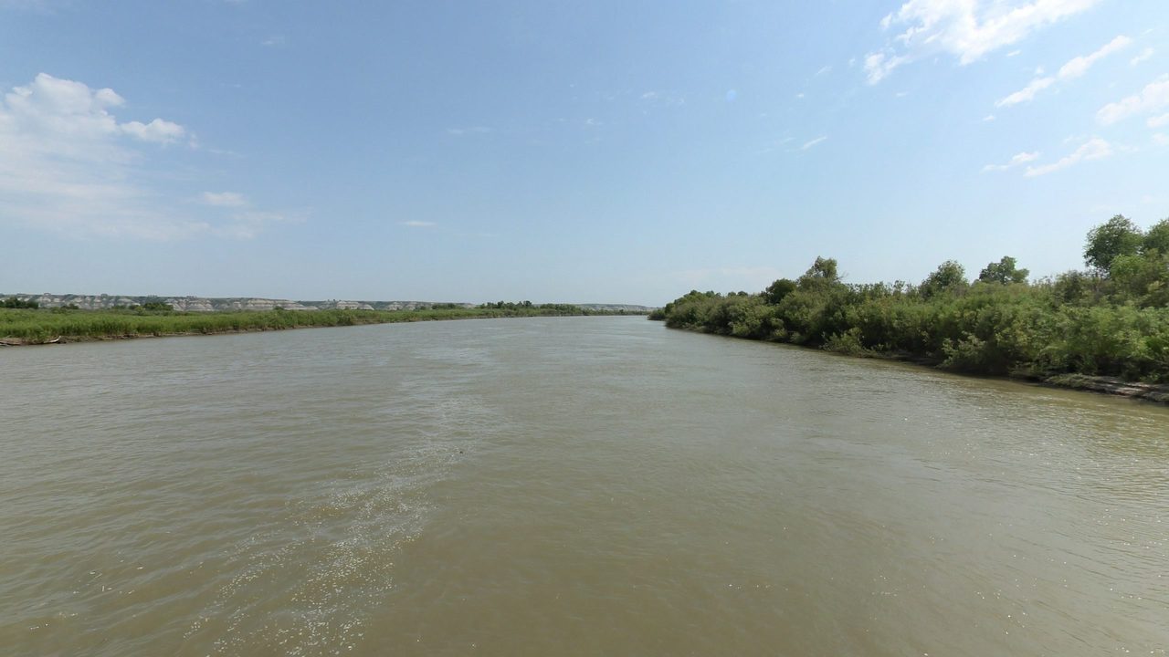 The Missouri River beside a brushy bank on the right, under a blue sky with light clouds, 2024