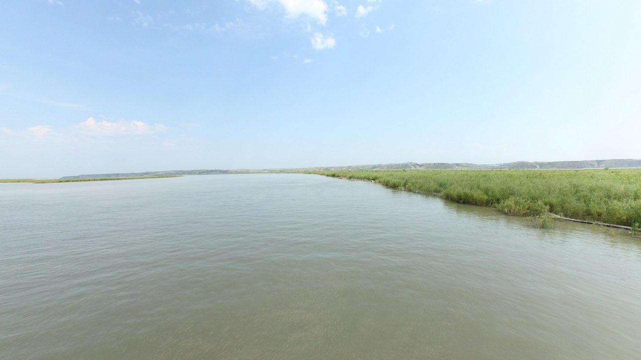The wide Missouri River beside a grassy bank on the right, under a pale sky, 2024
