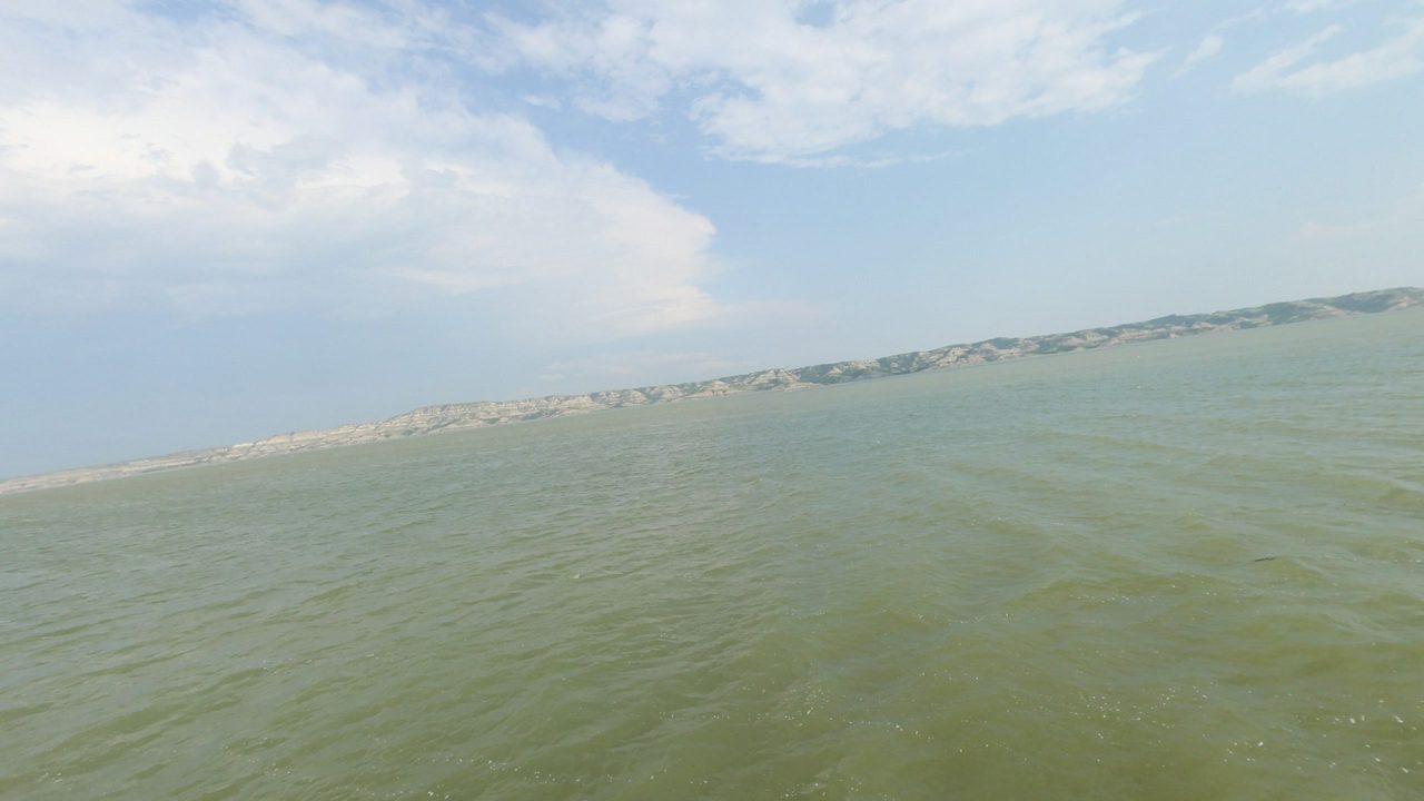 The wide Missouri River under a cloudy sky, with pale badland bluffs along the far shore, 2024