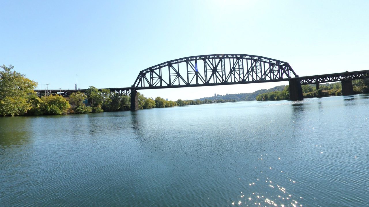 A steel truss railroad bridge across the Ohio River, with wooded banks under a clear blue sky, 2024