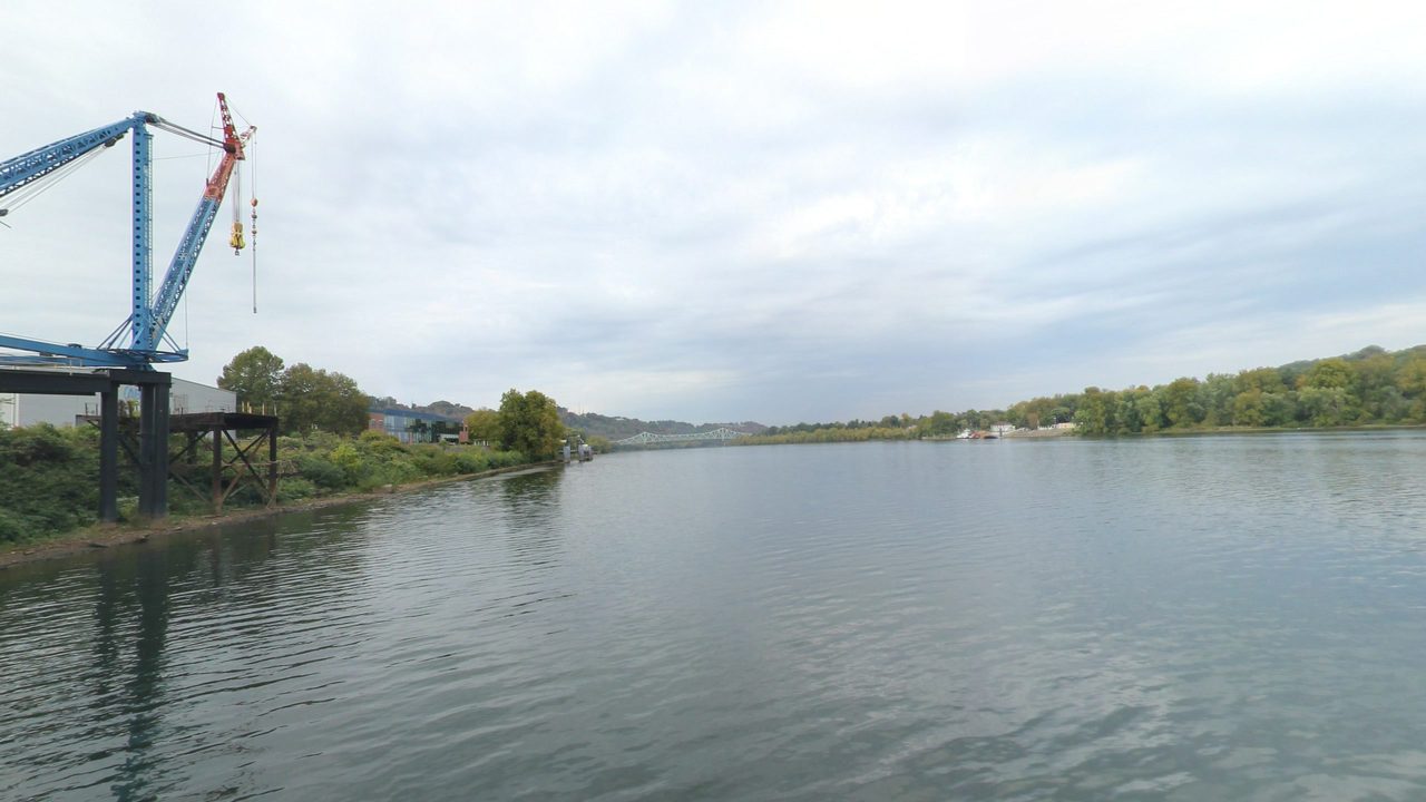 The Ohio River with a tall blue crane on an industrial dock on the left bank, and wooded shores and a distant bridge downstream, 2024
