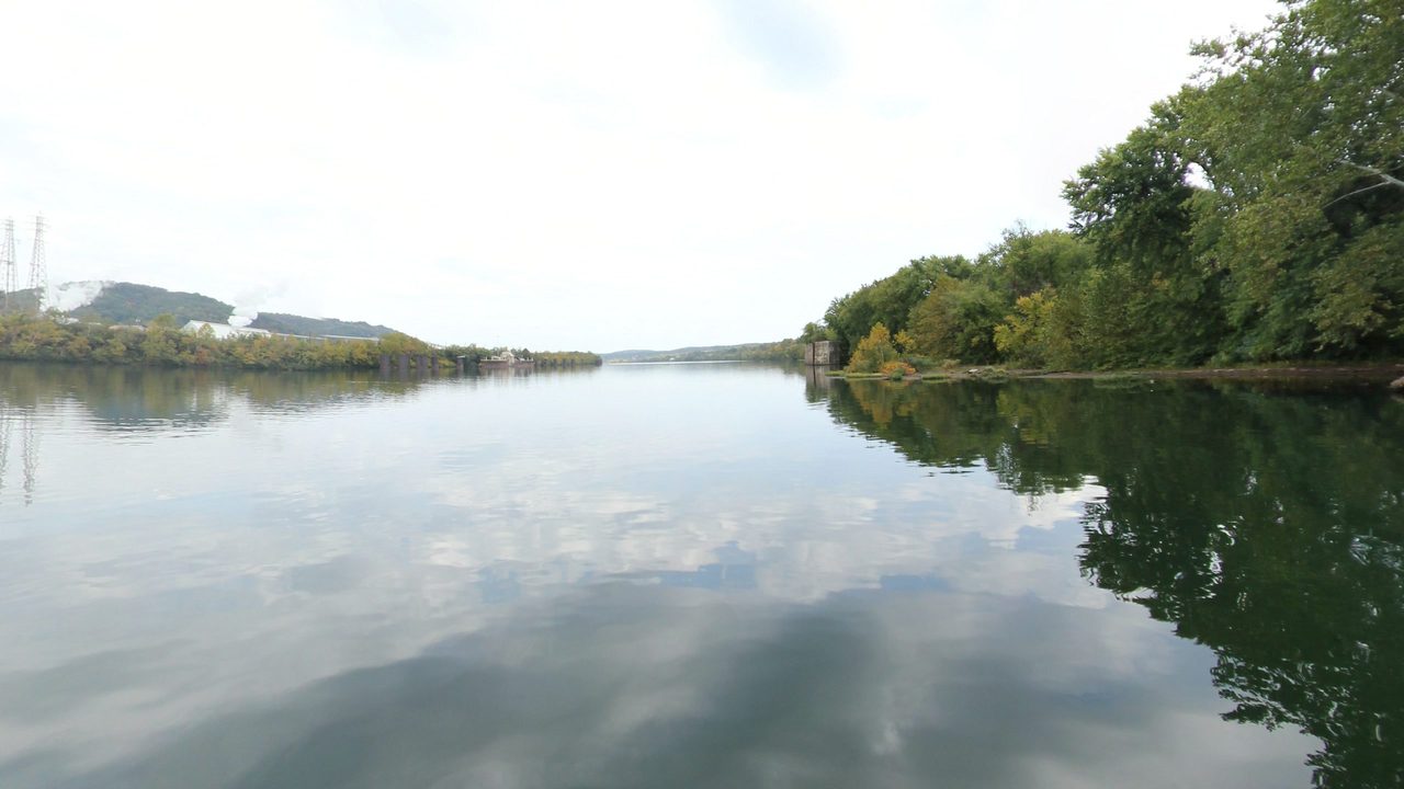 The calm Ohio River reflecting a cloudy sky, with a wooded bank on the right and low hills in the distance, 2024