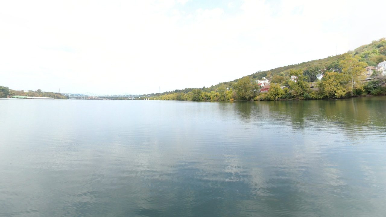 The calm Ohio River below a wooded hillside with houses on the right, under a white sky, 2024