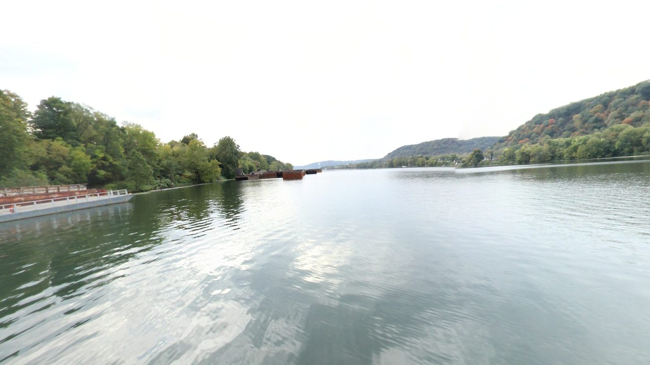 The wide Ohio River under a white sky, with a moored barge below wooded hills on the left, 2024