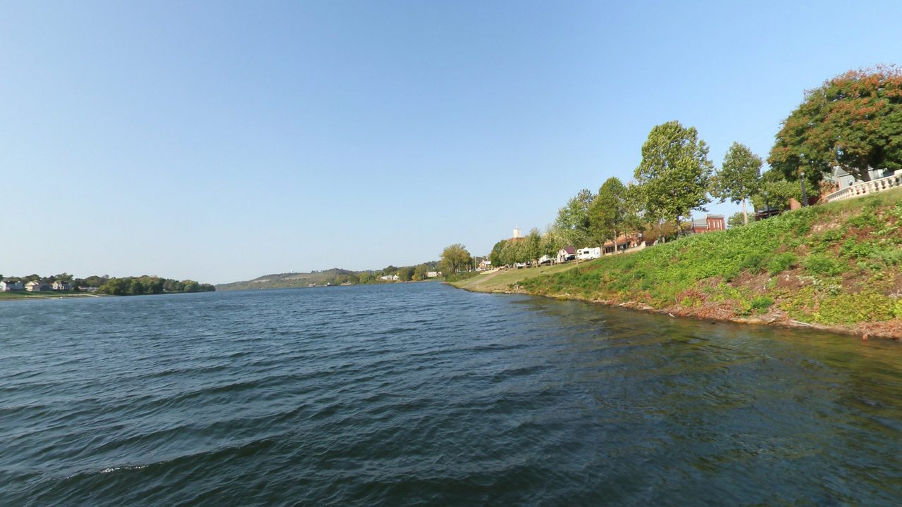 The Ohio River beside a steep green bank lined with trees and houses, 2024