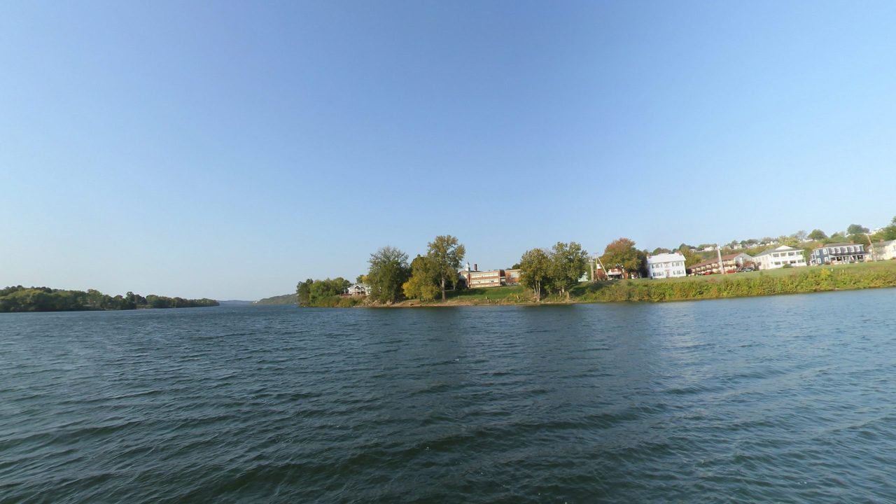 The Ohio River toward a wooded point and houses along the far shore, under a clear blue sky, 2024