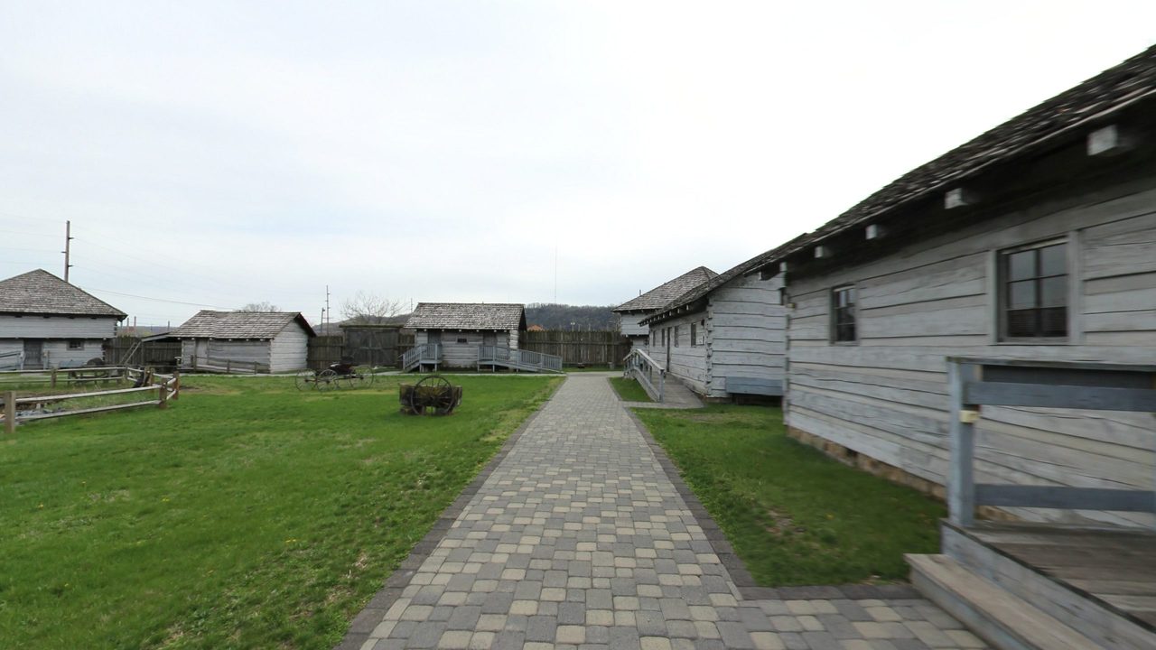 A brick path beside a long log building, with shingle-roofed log houses and a plank stockade across a lawn, 2025