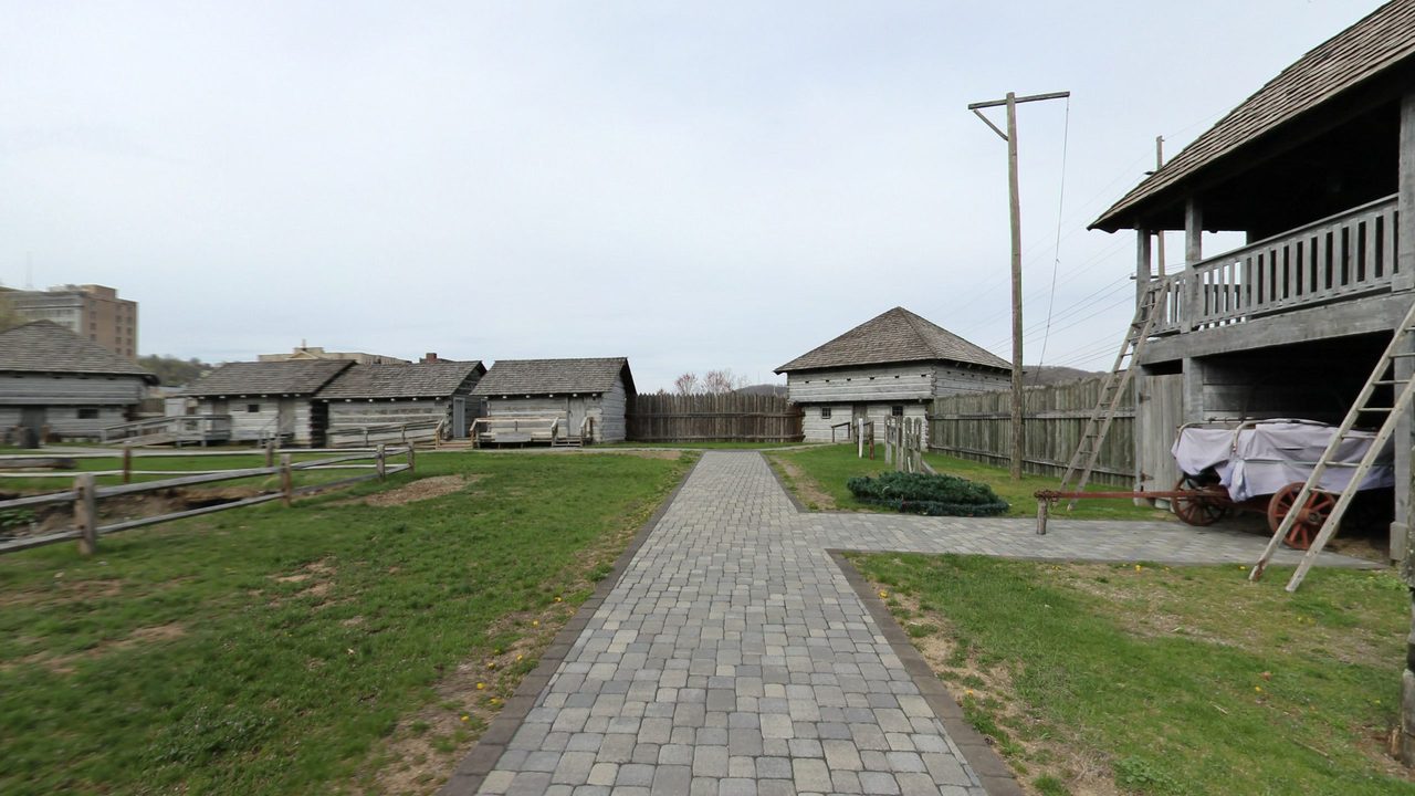 Reconstructed log blockhouses and a palisade around a lawn crossed by a brick path, with a covered wagon beneath a two-story blockhouse, 2025