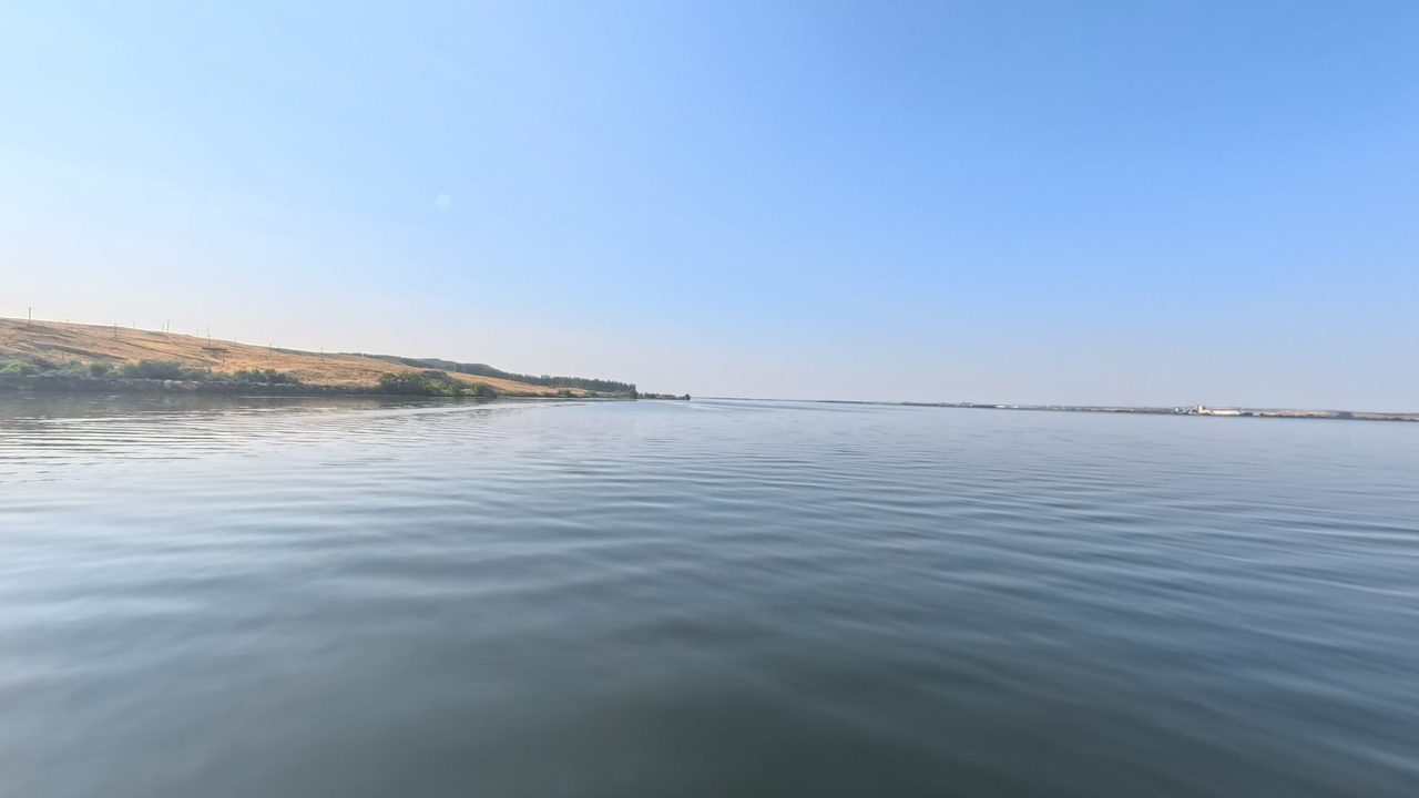 The calm Columbia River under a pale blue sky, with a low bluff on the left, 2025