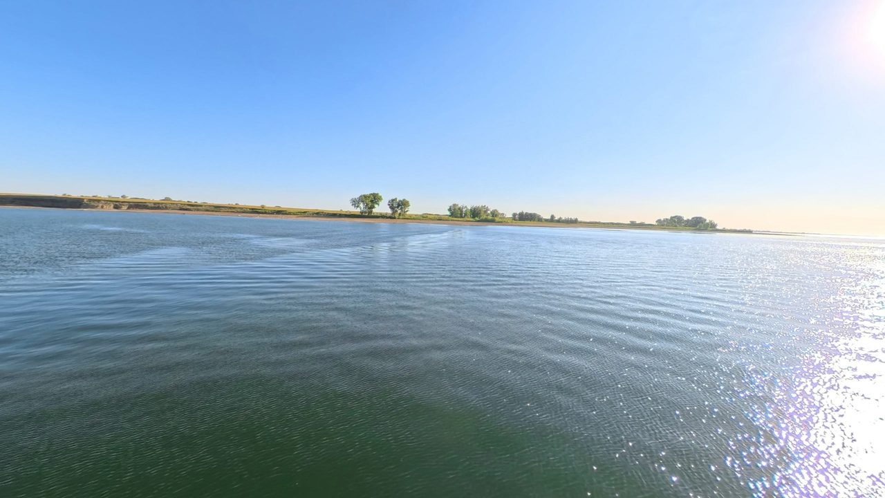 Lake Sakakawea rippling below a low sandy bluff and a line of trees, with sunlight glinting on the water, 2025