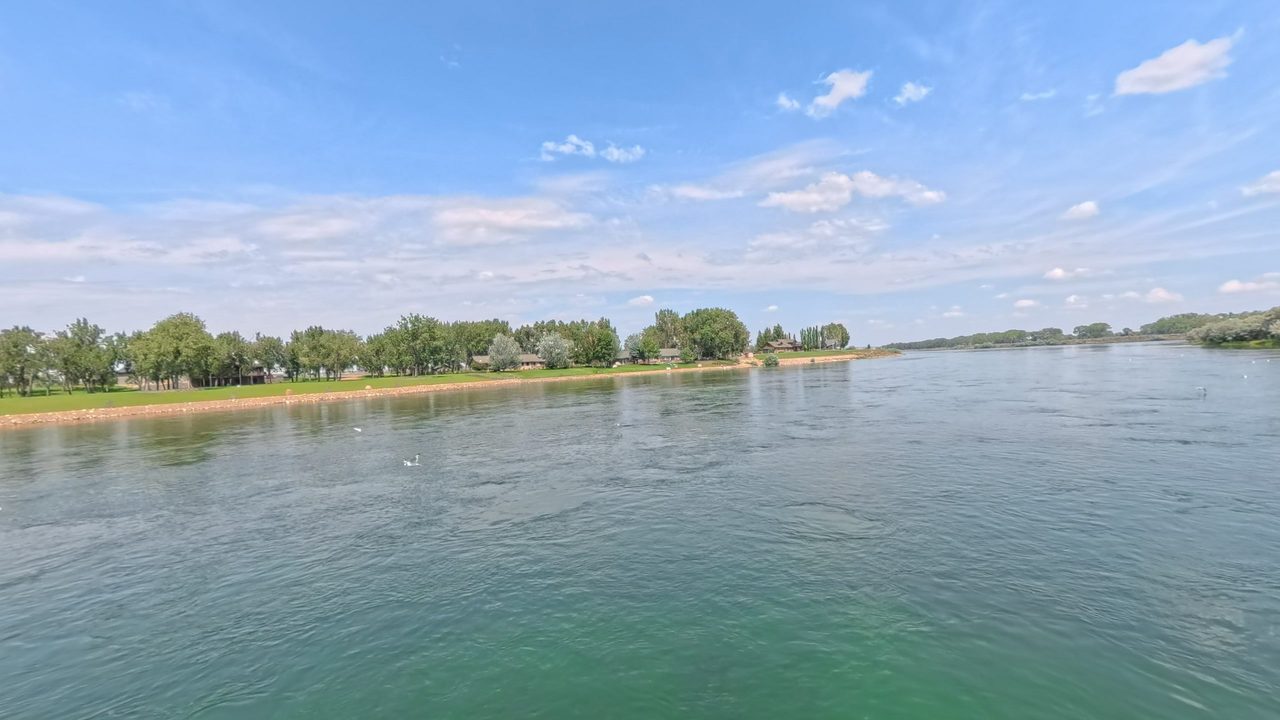 The Missouri River in clear green water, with a mown lawn and houses among trees on the left bank, 2025