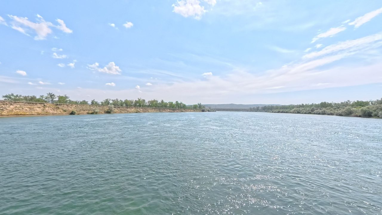 The Missouri River glinting under a blue sky with wispy clouds, between low wooded banks, 2025