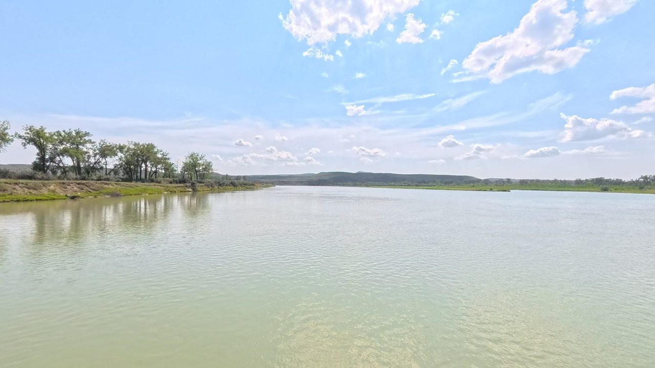 The calm Missouri River in pale green water, with cottonwoods on the left bank under scattered clouds, 2025