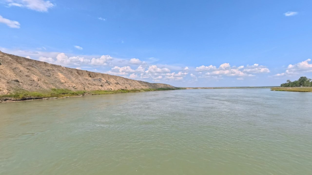 The Missouri River below a long grassy bluff under scattered white clouds, 2025