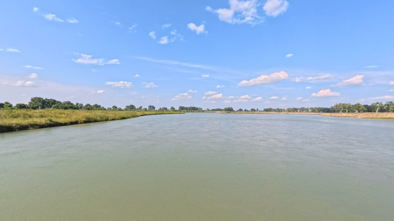 The Missouri River between grassy banks and a line of cottonwoods under scattered clouds, 2025