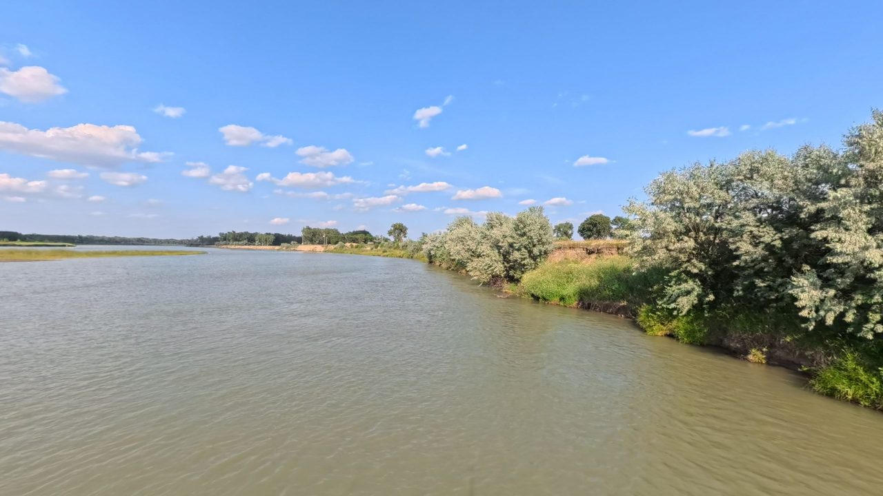 The Missouri River beside a bushy bank of silvery shrubs on the right, under a blue sky with small clouds, 2025