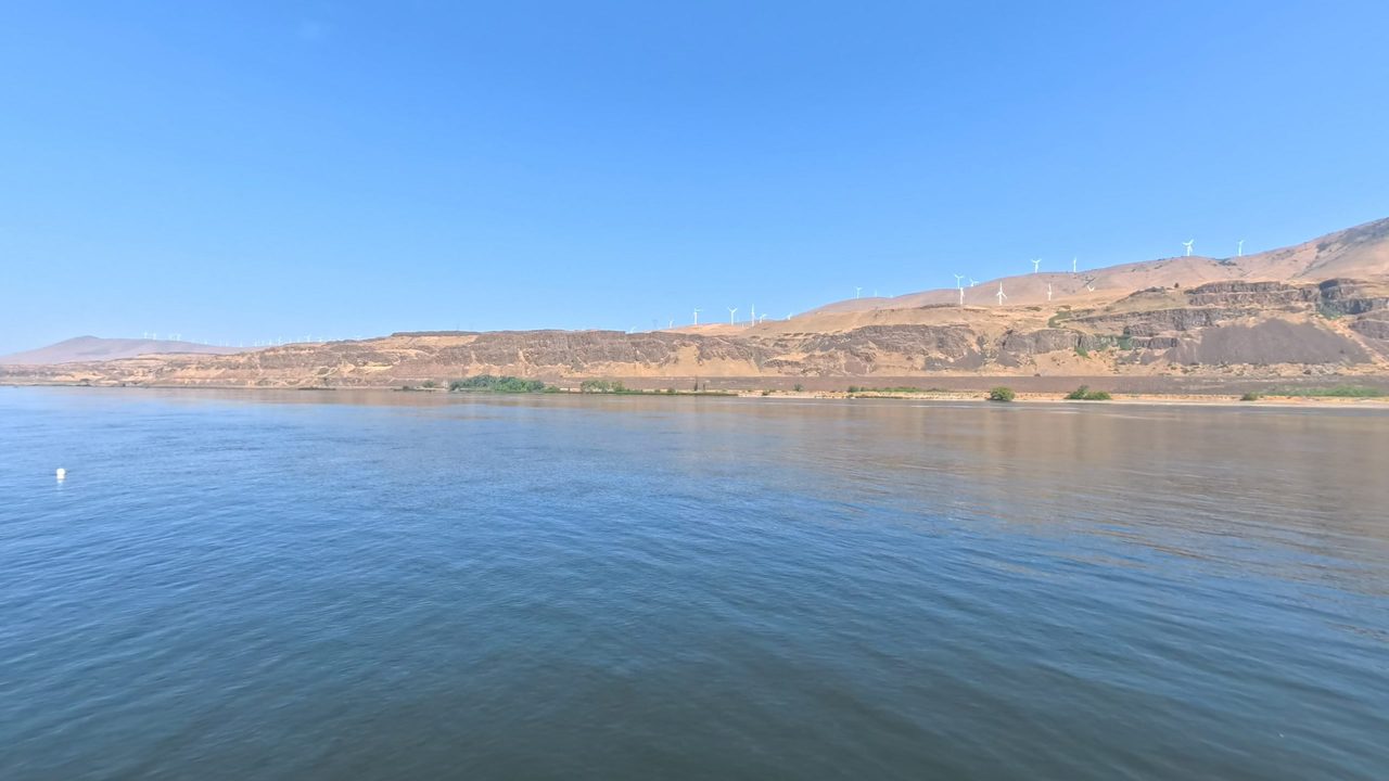 The calm Columbia River below dry hills and basalt cliffs, with wind turbines along the ridge, 2025