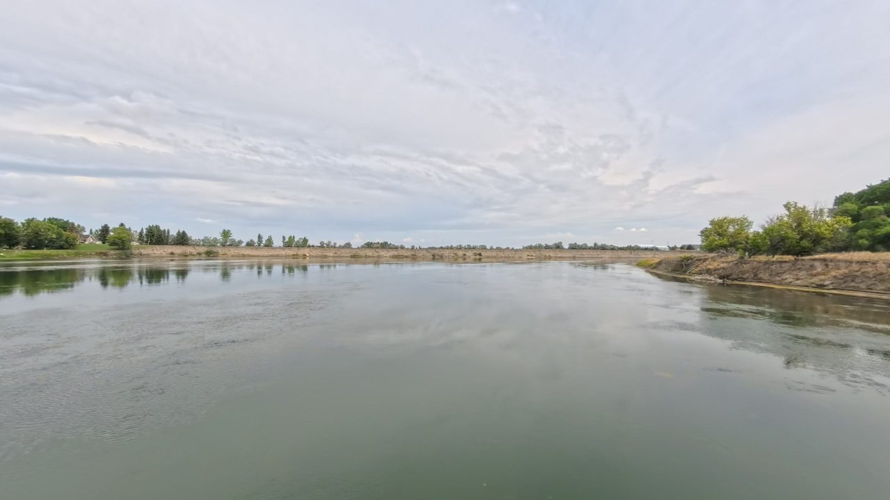 The calm Missouri River below a long grassy bank, with a house among trees on the left, under a hazy sky, 2025