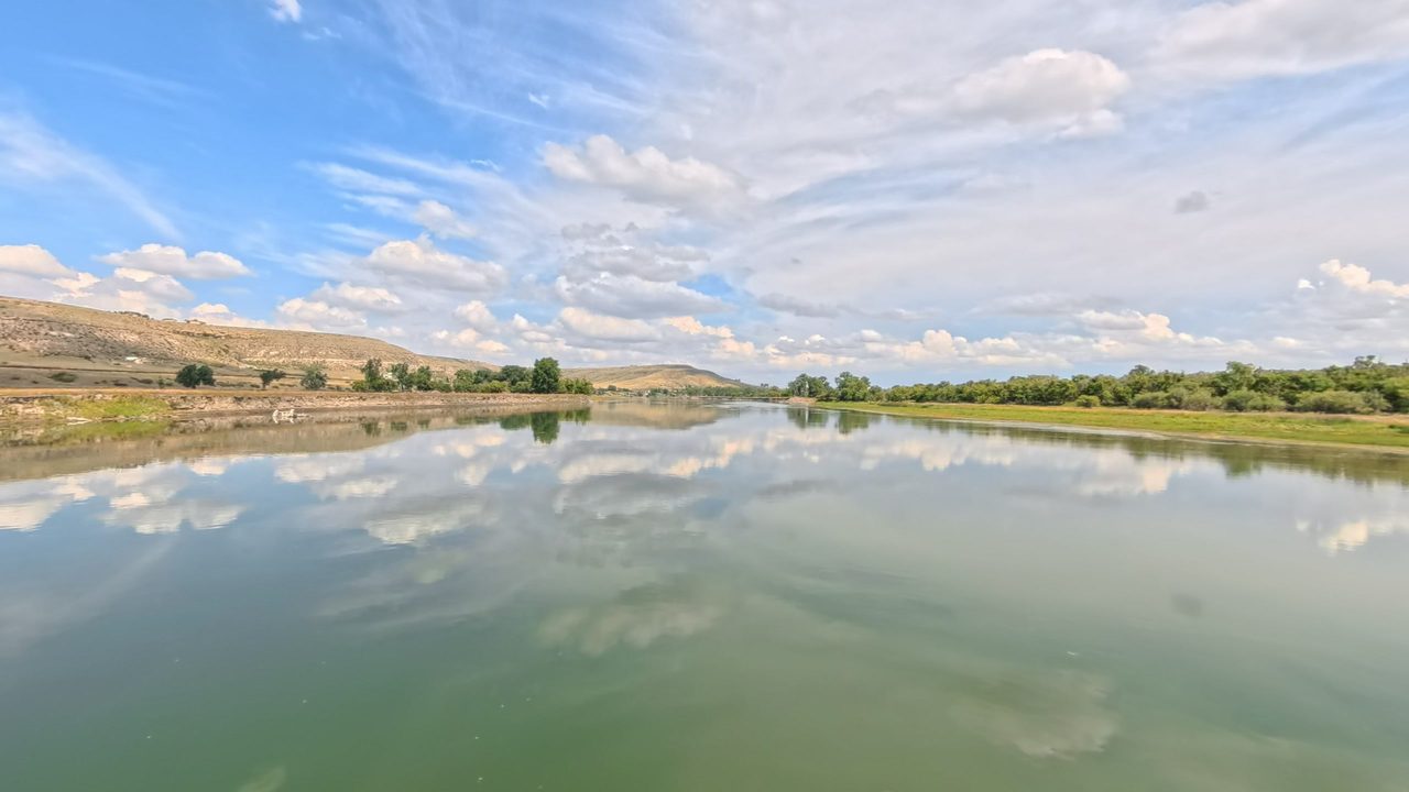 The calm Missouri River reflecting white clouds, with low grassy banks and hills beyond, 2025