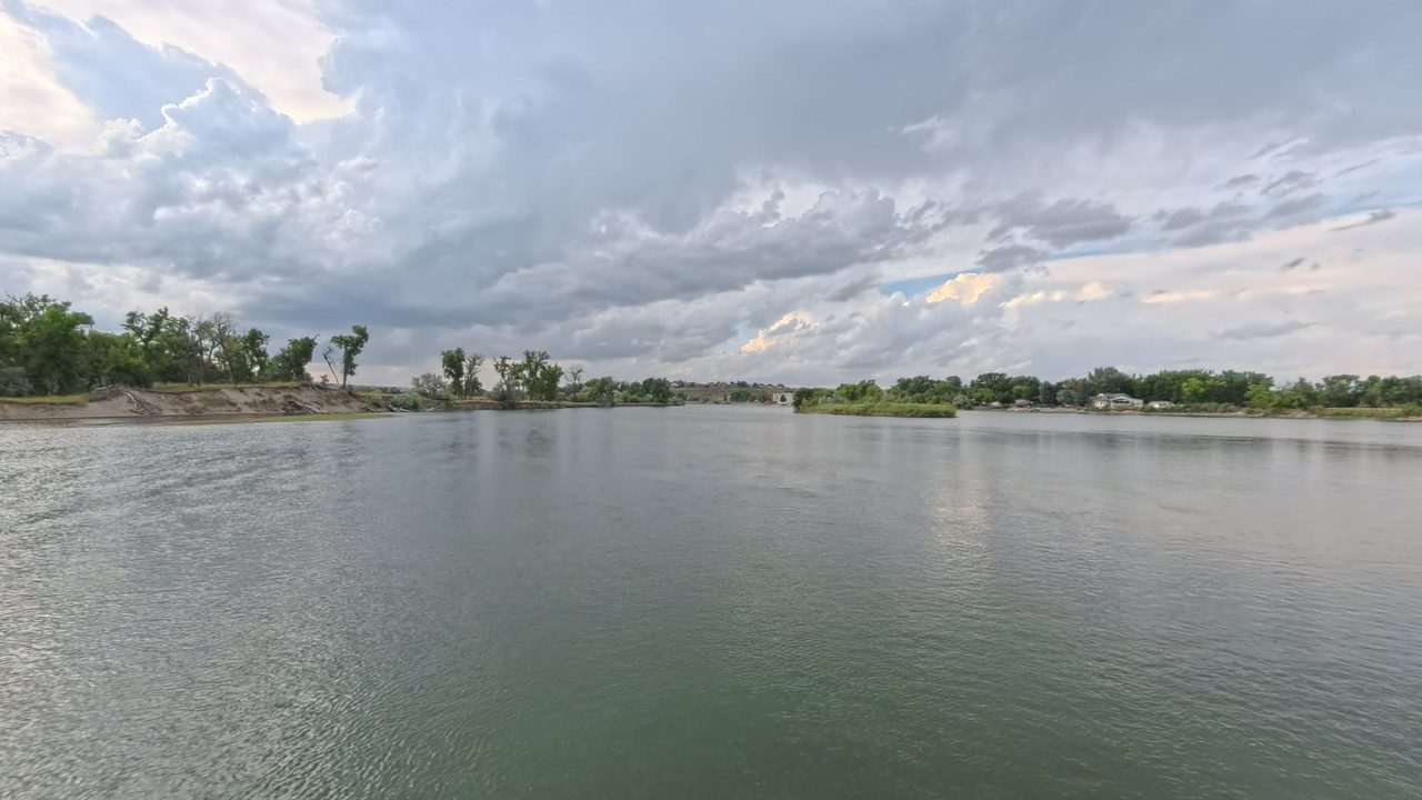 The Missouri River under heavy grey clouds, with leafy banks and riverside houses on the far right shore, 2025