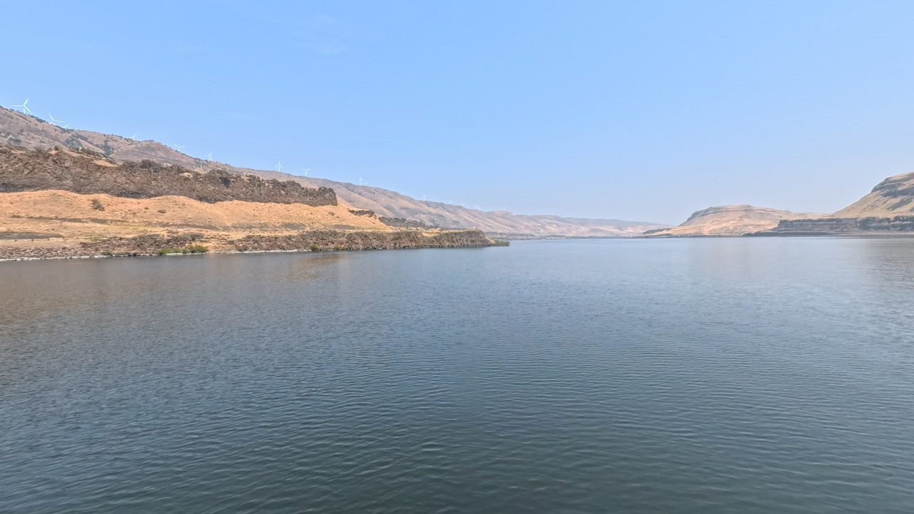The calm Columbia River below dry, grassy hills and dark basalt rimrock, 2025