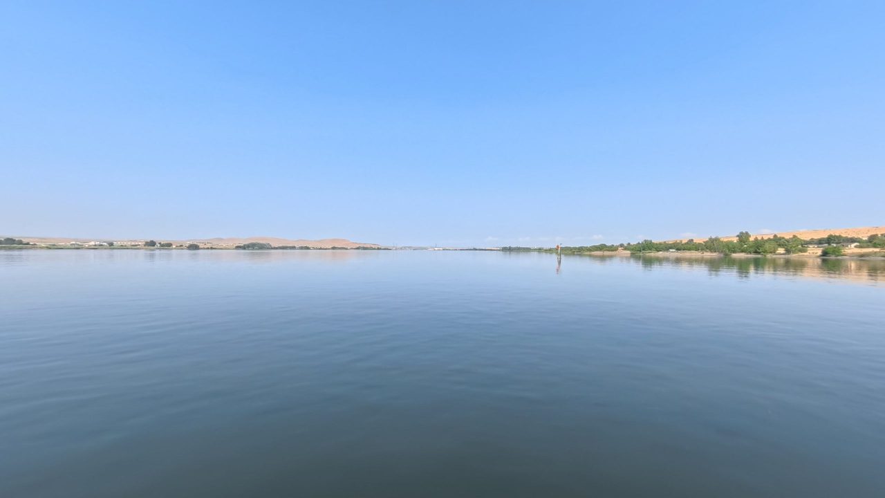 The calm Columbia River under a clear sky, with low dry hills and trees on the far shores, 2025