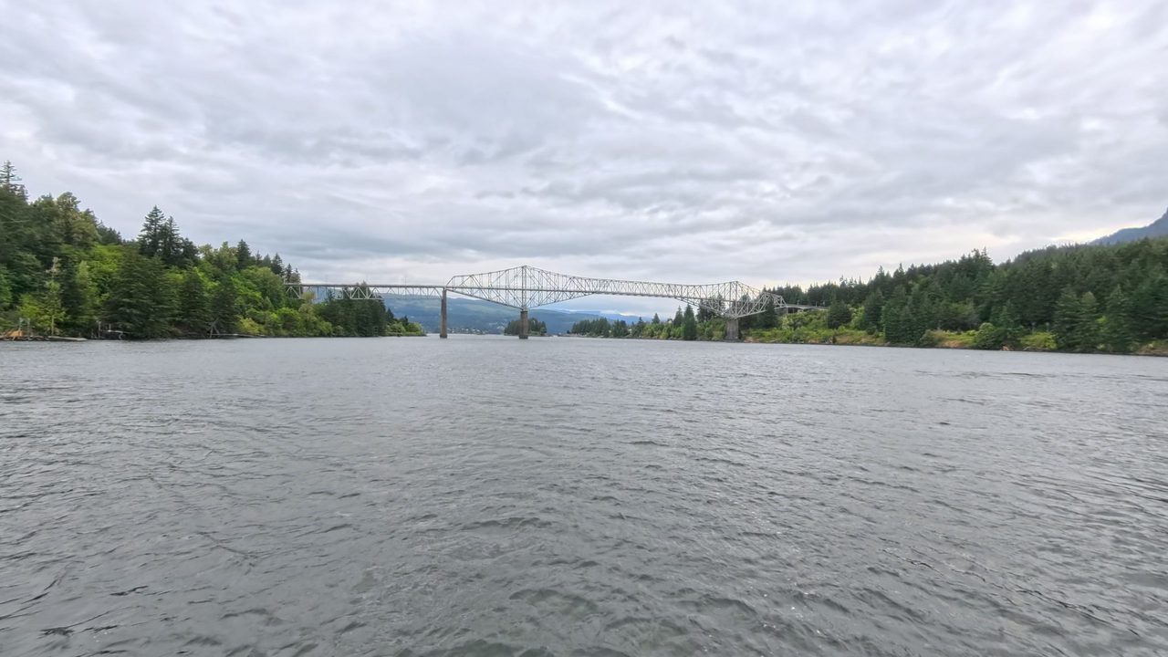 The Columbia River toward a long steel truss bridge between forested shores, under a cloudy sky, 2025