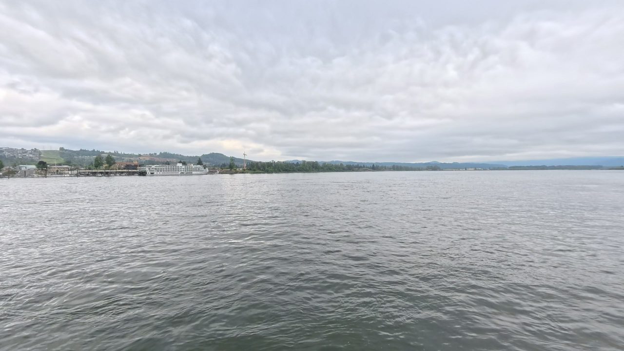 The Columbia River under a mottled grey sky, with a white riverboat moored at a small town's waterfront, 2025