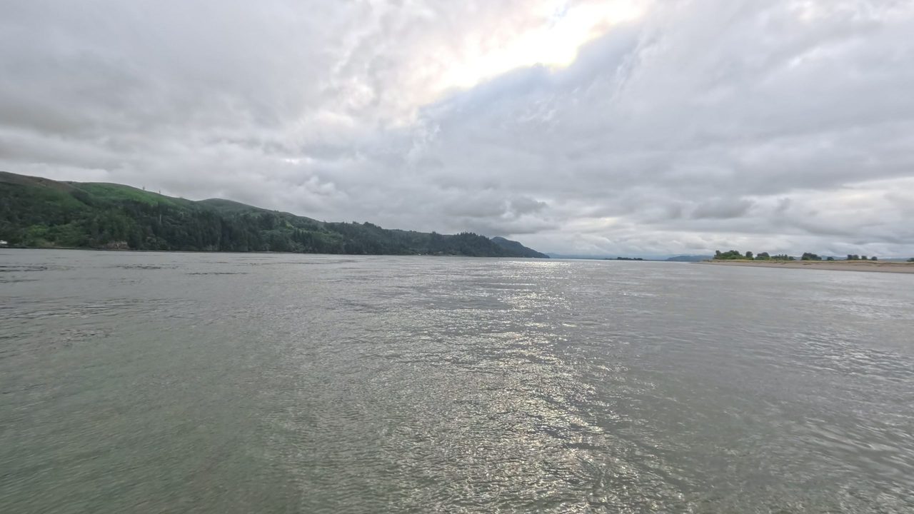 The wide Columbia River under heavy clouds, with forested hills along the left shore, 2025