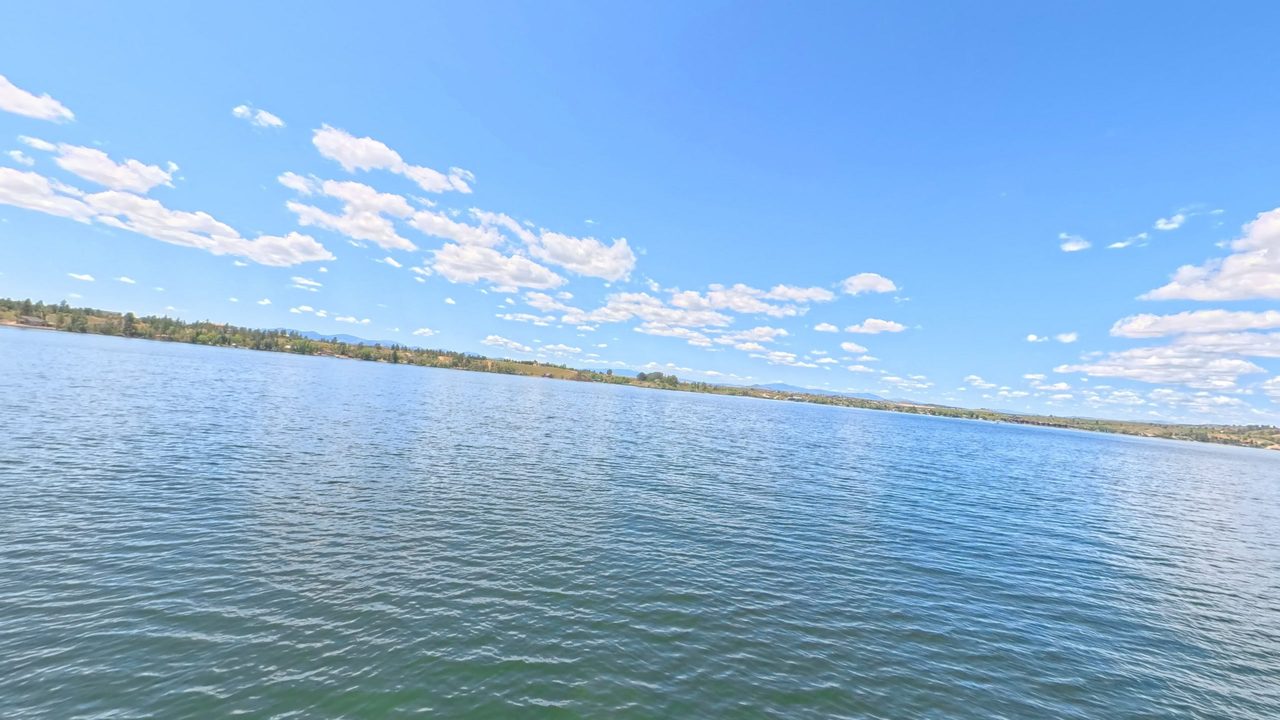 Canyon Ferry Lake under a blue sky with scattered clouds, with a wooded shore dotted with houses, 2025