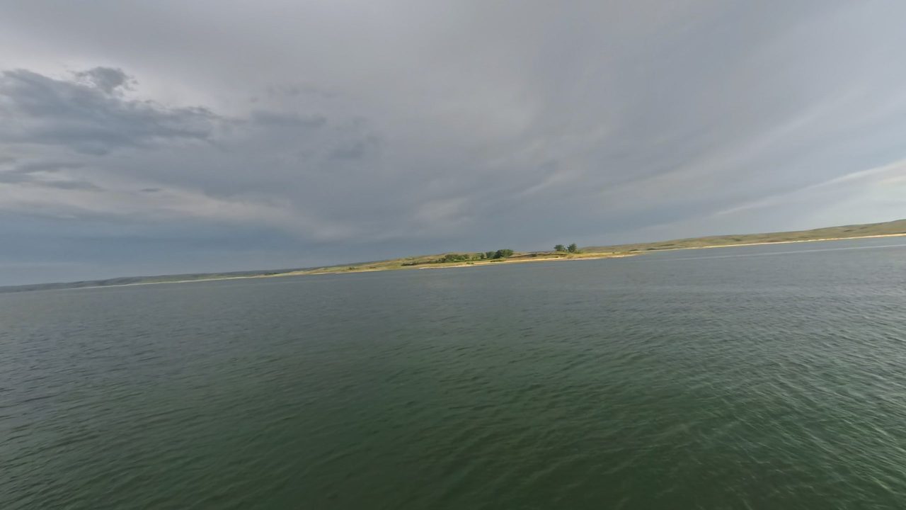Lake Oahe under a dark, cloudy sky, with a low grassy point across the water, 2025