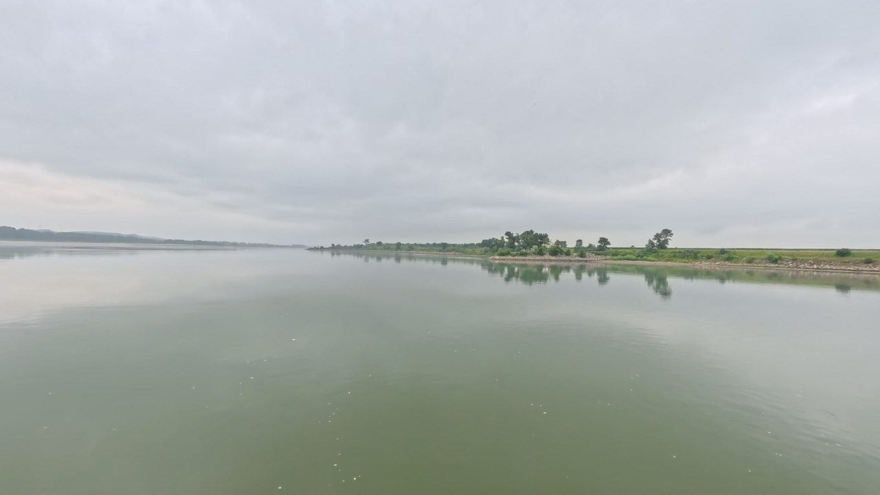 The calm Missouri River with a low wooded point in the distance, under an overcast sky, 2025