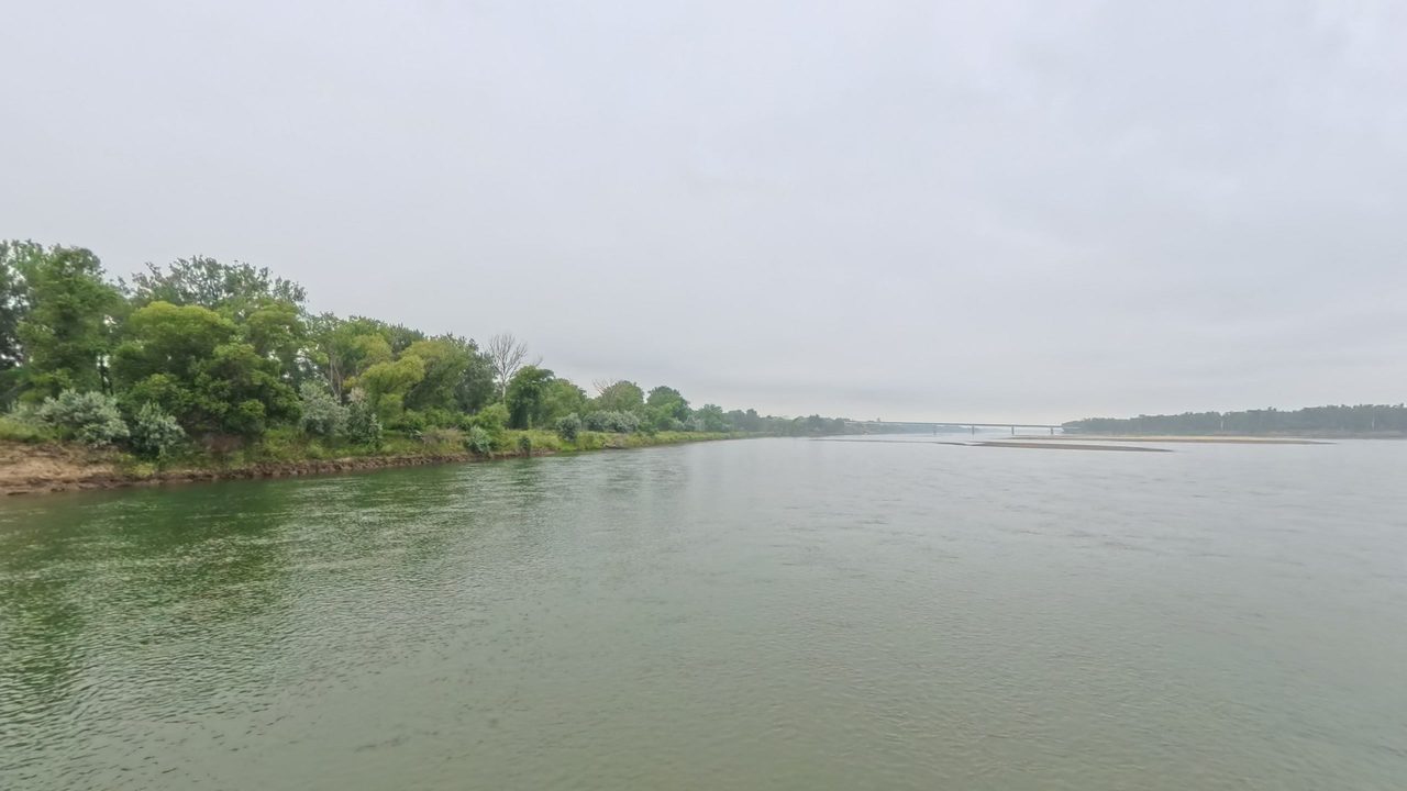 The Missouri River under a hazy, overcast sky, with a leafy bank on the left and a long bridge in the distance, 2025