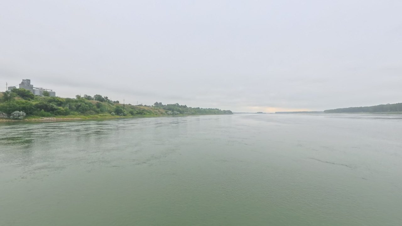 The calm Missouri River below a grassy, shrubby bank with grain silos at the top on the left, under a hazy sky, 2025