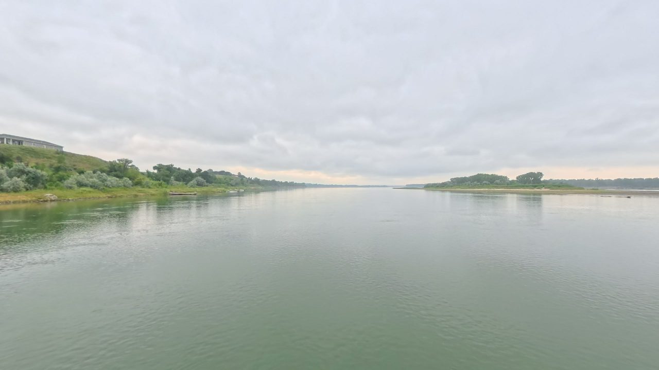 The Missouri River beside a grassy bank topped by a building on the left, with a wooded island downstream, 2025